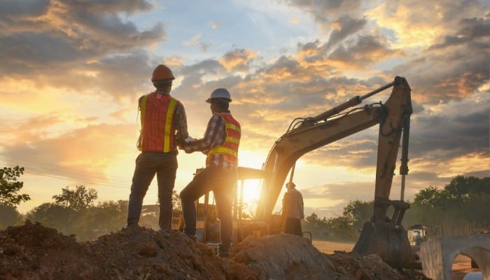 Two men in plaid shirts and orange high-visibility vests stand on a mound of dirt next to an excavator.