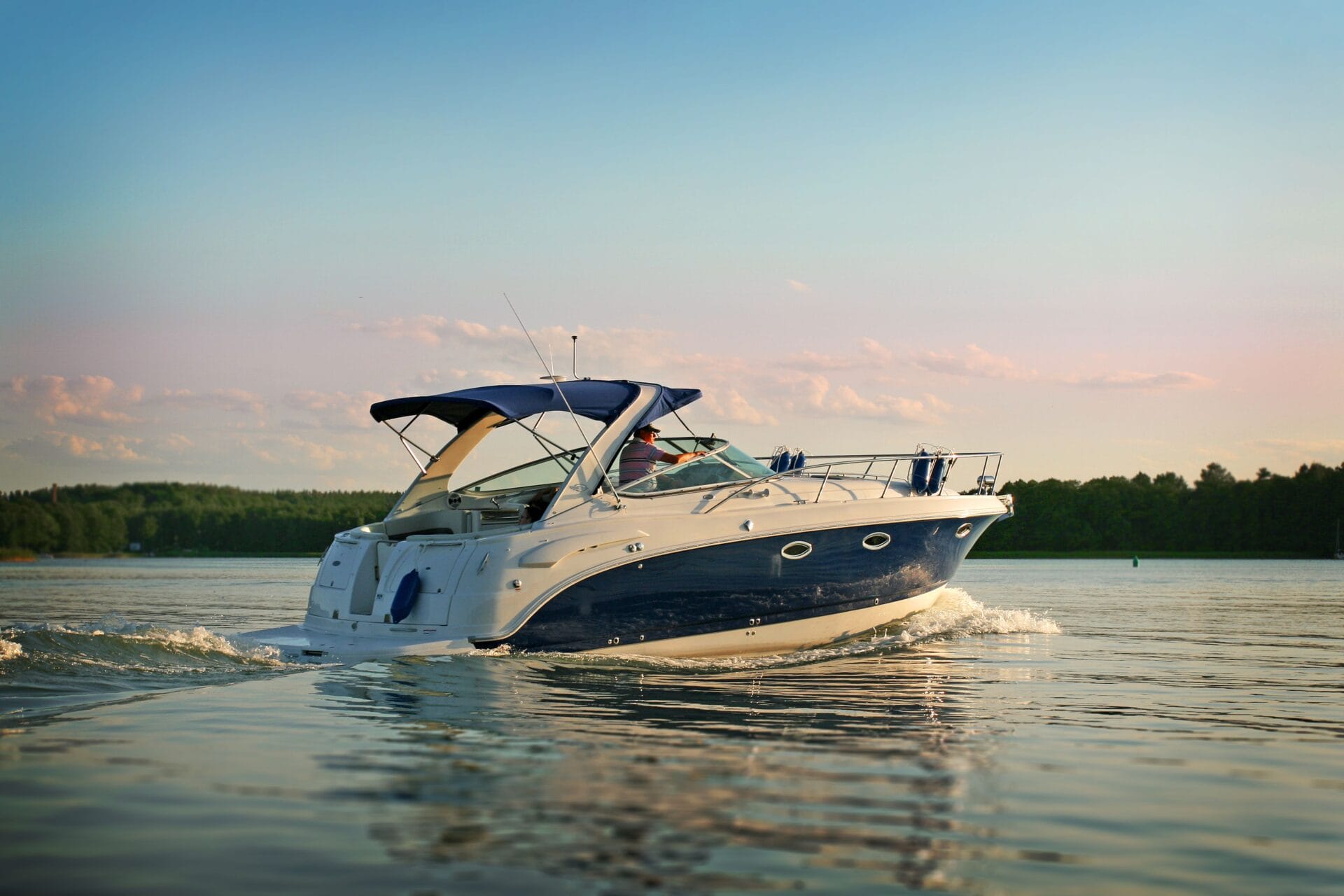 A person is driving a white and blue yacht boat by himself during the day out on a lake surrounded by trees.
