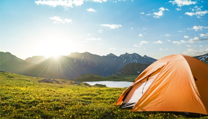A small, orange tent pitched in a meadow with yellow flowers next to some water and mountains on a sunny day.