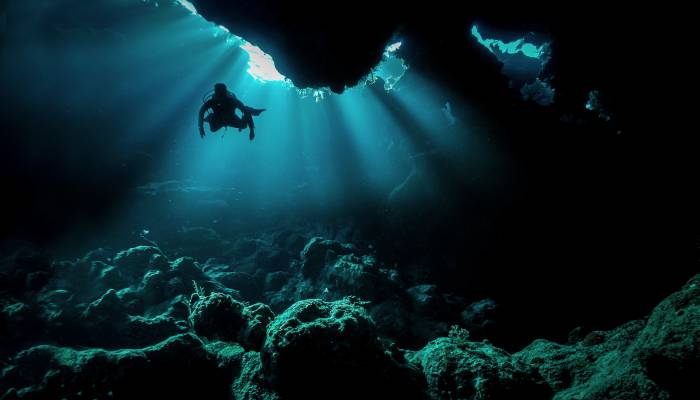 A lone scuba diver navigating through a dark, rocky reef terrain and swimming in advanced diving conditions.