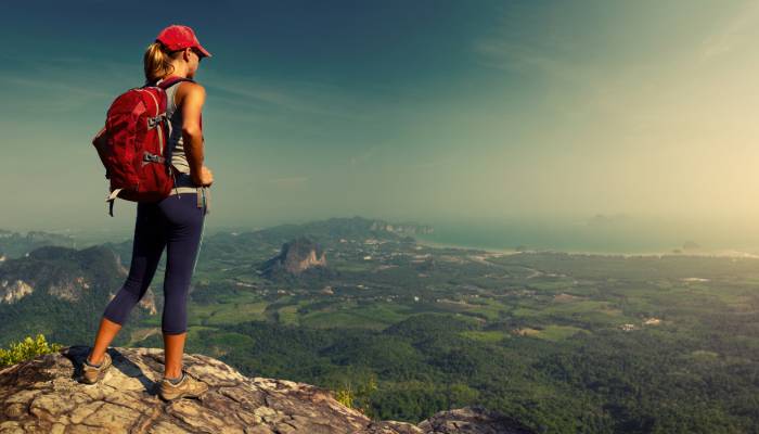 A woman wearing a red backpack and hiking clothes stands on a rock overlooking a beautiful view of trees and rocks.