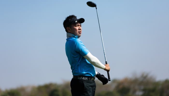 A professional golf player in a blue polo and white undershirt staring into the distance as he completes a swing.