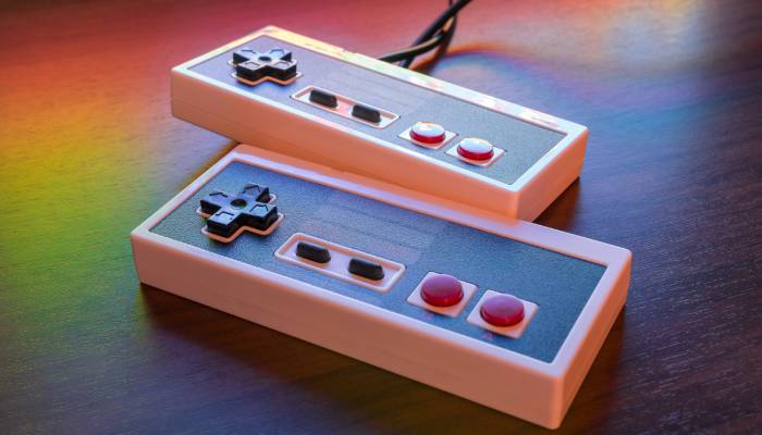 A close-up view shows two retro controllers with tangled cords sitting on a wooden surface. A rainbow appears over them.