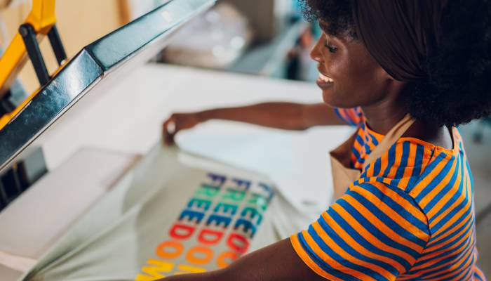 A textile worker, wearing a beige apron, placing a graphic t-shirt with a rainbow design onto a heat press.