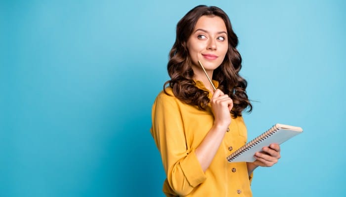 A young woman is holding a notebook and pen. She's holding the pen near her face and is thinking about something.
