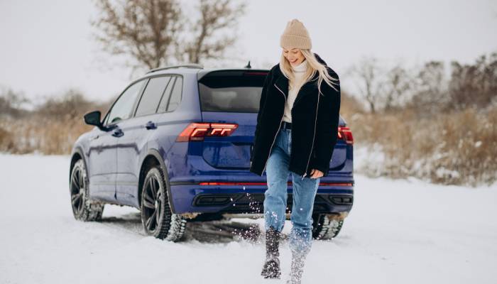 A smiling woman wears a black coat, jeans, a tan hat, and a white sweater as she kicks up snow. A blue car is in the background.