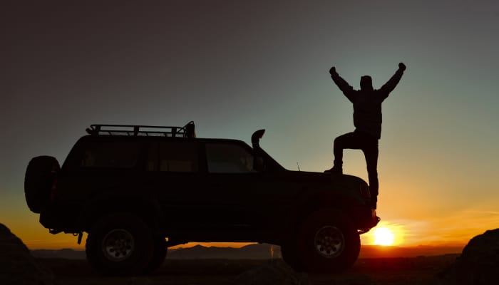 The silhouette of a man standing on the front bumper of his off-roading vehicle with an orange setting sun in the background.