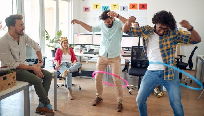 A group of employees in their office with two employees hula hopping and two other employees sitting and smiling.
