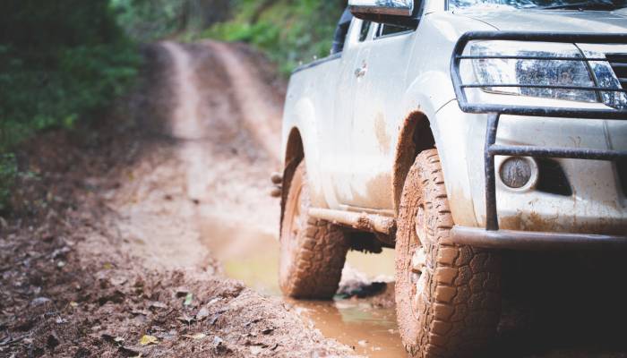A light gray truck with very muddy wheels drives down a dirt path with puddles and fallen leaves on it.