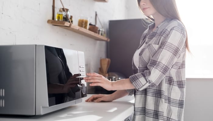 A young woman wearing a plaid shirt is standing by her kitchen counter and using her silver and black microwave.