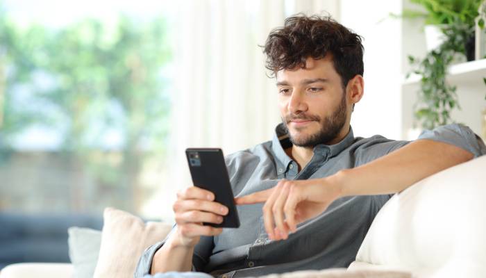 A man is sitting on a white couch in a brightly lit room. He is using his finger to scroll on his cell phone.
