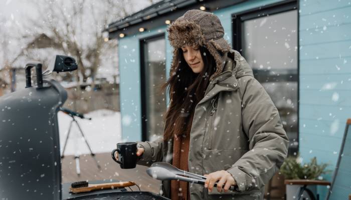 A woman wearing a winter coat and hat is holding a mug and a pair of tongs while grilling in the snow.