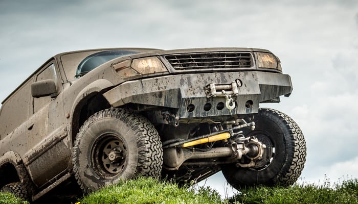 On a grassy hill beneath a cloudy sky, a dirty off-road truck sits with its headlights on and its tires slightly turned.