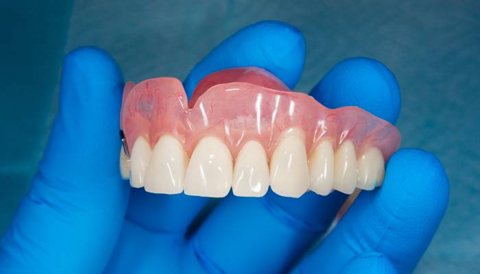 A close-up of an individual wearing a blue medical glove holding a human denture of the upper jaw on a blue background.