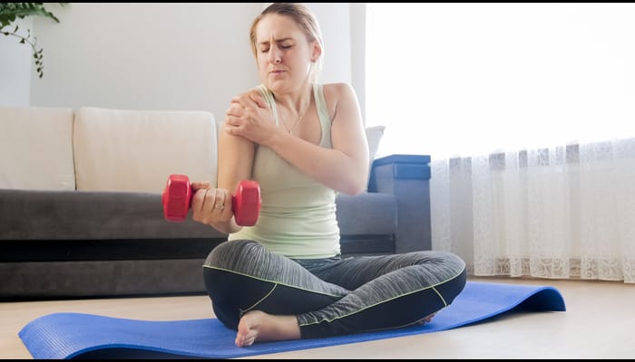 A woman sitting on a yoga mat holds a pink dumbbell in one hand while gripping her shoulder in pain with her other hand.