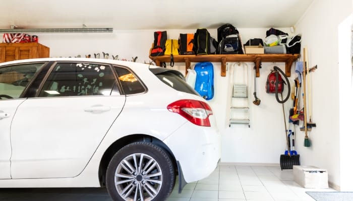 A white van parked in a garage featuring white walls, tiled floors, and wooden shelves on the wall filled with clutter.