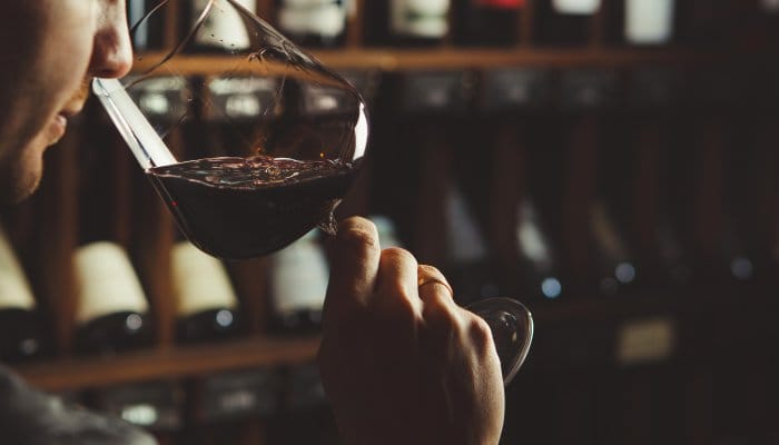 A close-up shot of a man in a wine cellar holding a glass of red wine up to his face, sniffing the contents.