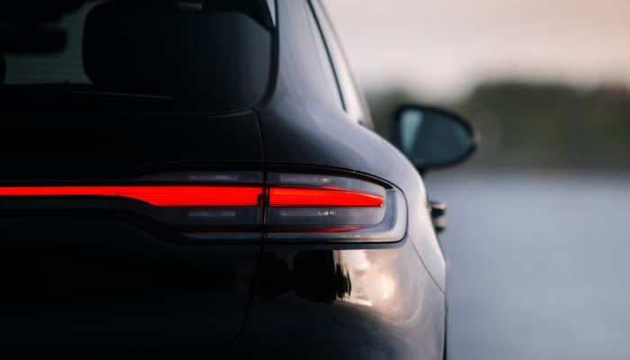 A close-up of the rear of a Porsche SUV in the evening. The Porsche's horizontal red tail light is activated.