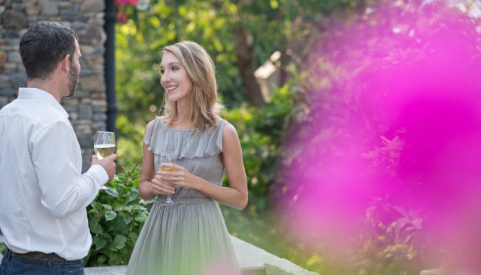 A woman in a gray dress holding a champagne glass smiles at a man in a white shirt and jeans.