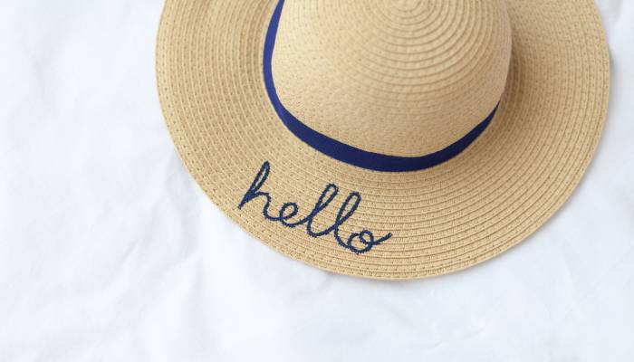 A woven hat with a blue ribbon and “hello” written on its brim in blue thread on a white background.