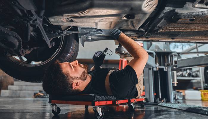 A young man in a black shirt lays on a garage creeper while inspecting the undercarriage of a vehicle with one hand.