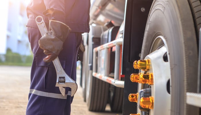 A close-up of a mechanic wearing gloves and holding a large wrench while standing next to the wheel of a semi-truck.