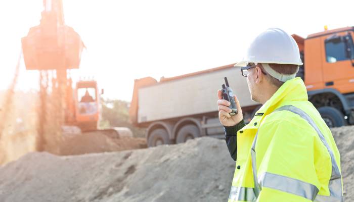 A man wearing a yellow safety vest and a white hard hat is holding a radio. He is standing on a construction site.