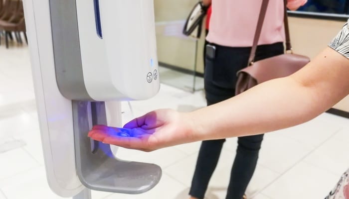 A person in a public space uses a touch-free hand sanitizer dispenser. The dispenser shines a blue light on their hand.