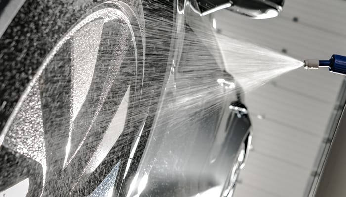 A close-up view shows a black vehicle's tire being sprayed with a liquid from a blue and white sprayer.