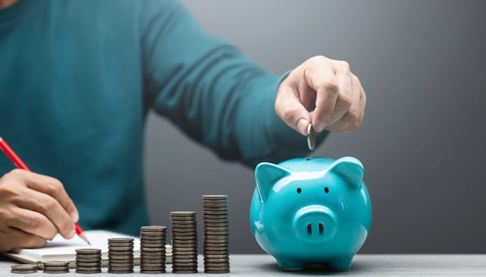 A man is dropping a coin into a blue piggy bank. There are several stacks of coins sitting next to the piggy bank.