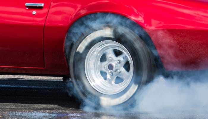 A red drag racing vehicle spins its wheels on the road. The wheels create white smoke.
