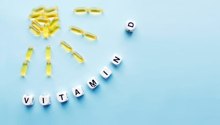 White blocks spell out “vitamin D” near a sun made out of vitamin D capsules on a blue background.