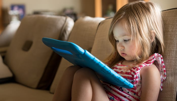 A young preschool girl sitting on the couch at home with an iPad on her lap. She's very focused on the screen.