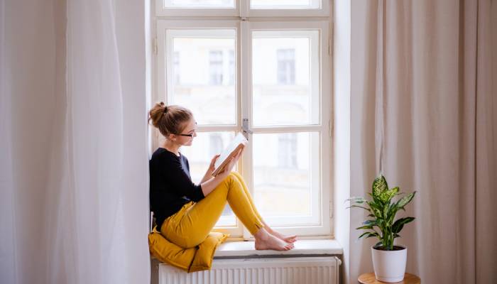 A woman in a black shirt, yellow pants, and black glasses sitting on a yellow pillow on a windowsill, reading a book.