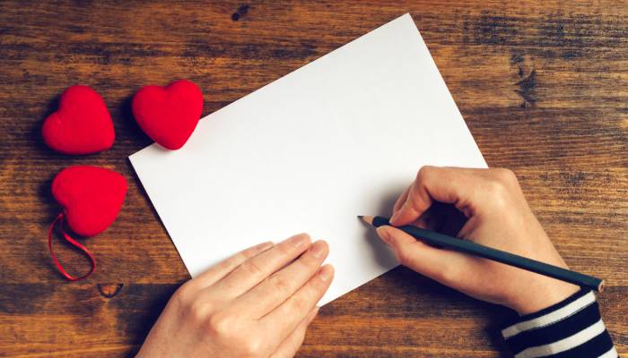Someone holds a pencil over a blank white sheet of paper. There are three red hearts on the table next to the paper.