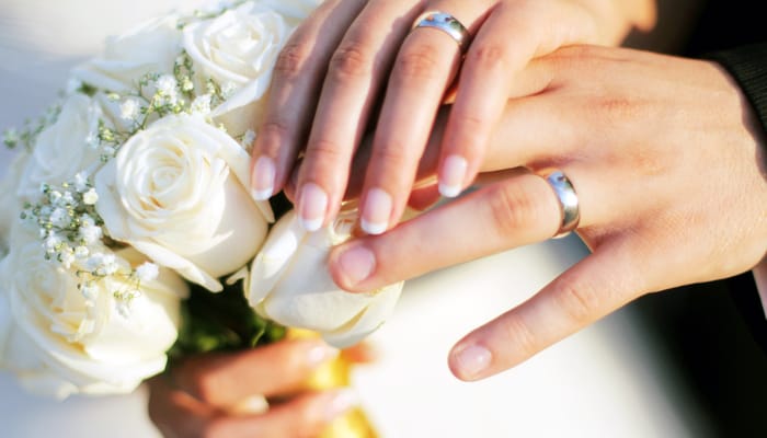 A bride and groom wearing matching silver wedding rings while the bride holds a bouquet of white roses.