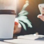 A person sitting at a picnic table with a coffee in a to-go cup with a black lid. They're holding a phone in the other hand.