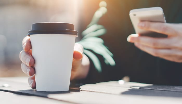 A person sitting at a picnic table with a coffee in a to-go cup with a black lid. They're holding a phone in the other hand.