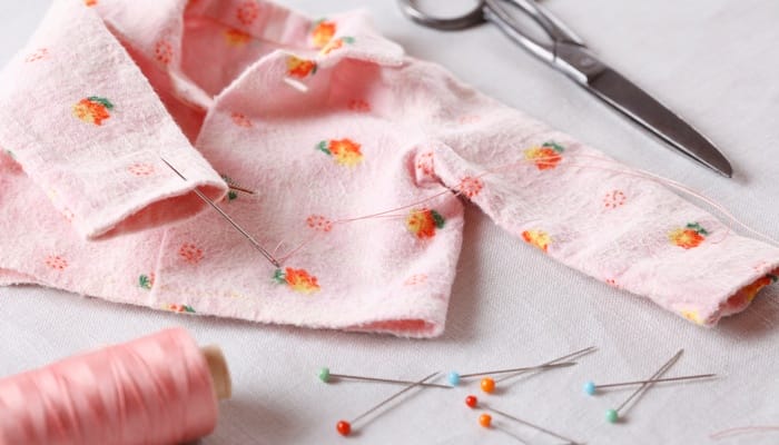 A handsewn pink baby jacket laying on a table with stick pins nearby and a pair of silver scissors next to a sleeve.
