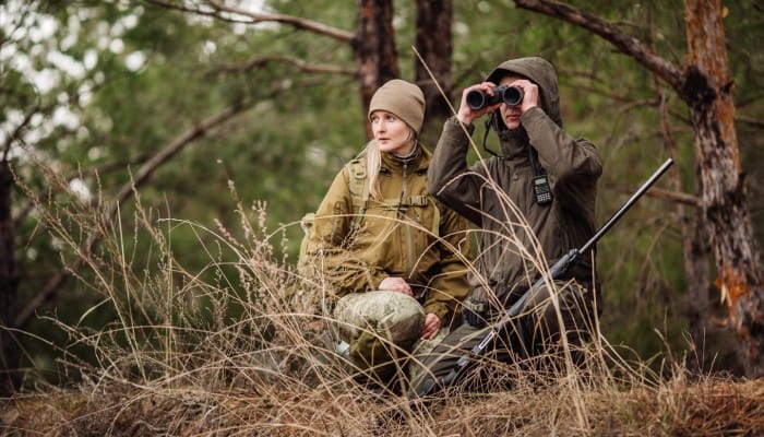 A woman wearing a beanie kneeling next to a man looking through binoculars and balancing a gun on his knee.