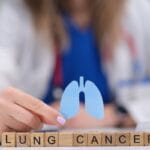 A doctor sits at her desk and holds a paper cutout of lungs over wooden blocks that spell out "Lung Cancer."