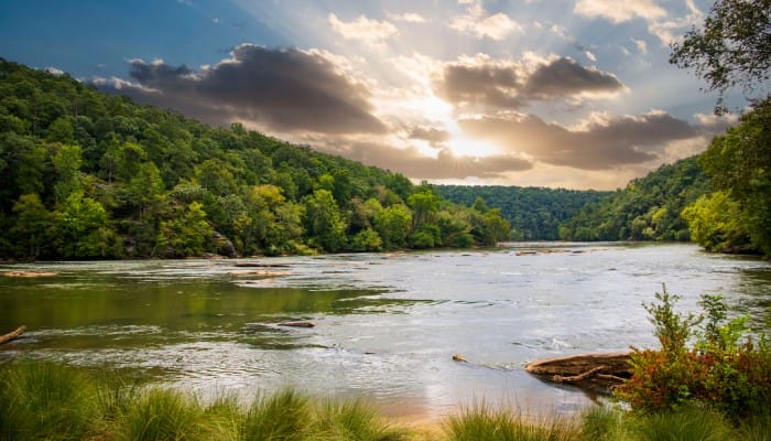 A wide river flows through a forested and mountainous region. The sun shines through the clouds in the sky.