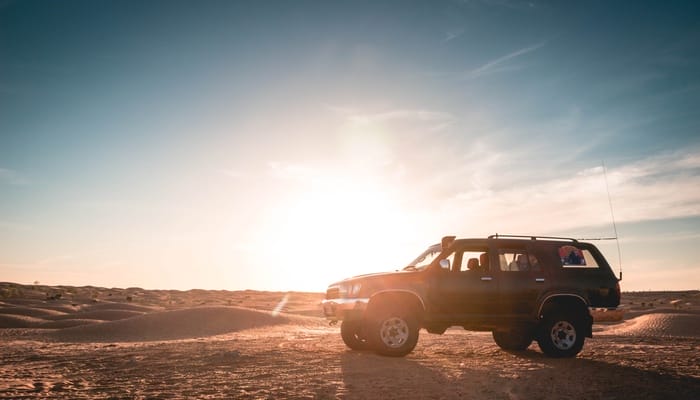 An off-roading vehicle packed with gear traverses a sandy landscape with the setting sun in the background.