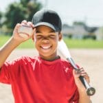 A smiling young boy wears a black baseball hat and a red shirt. He is holding a baseball and resting a bat on his shoulder.