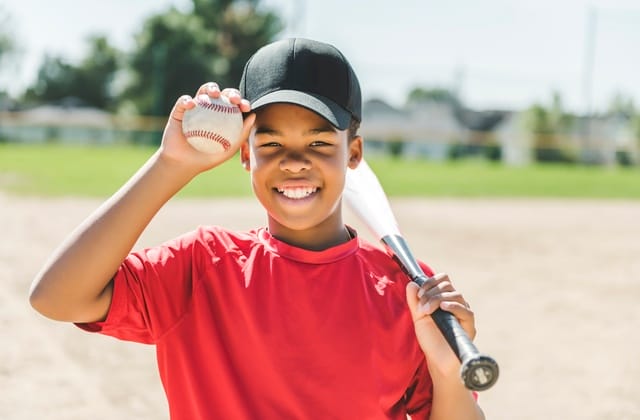 A smiling young boy wears a black baseball hat and a red shirt. He is holding a baseball and resting a bat on his shoulder.