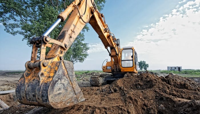 The bucket of an orange excavator is digging dirt to create a large hole with a mature tree in the background.