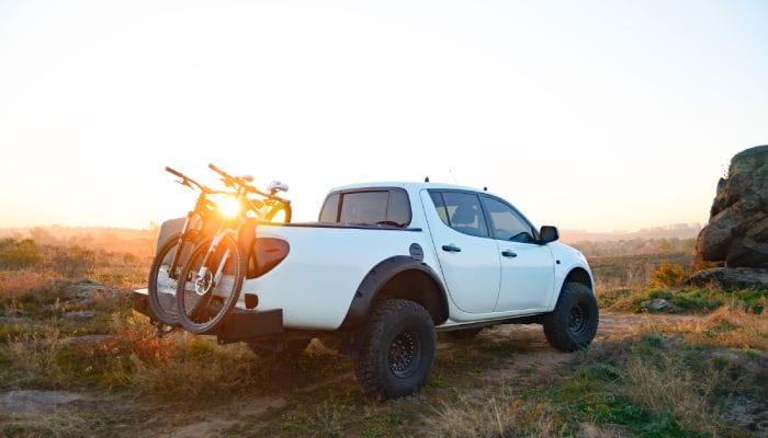 A white pickup truck with two bikes on the back of the trunk drives along an off-road mountain path as the sun sets.