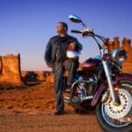 A motorcycle rider parked near the edge of the mountains. His helmet is on his knee as he rests against his bike.