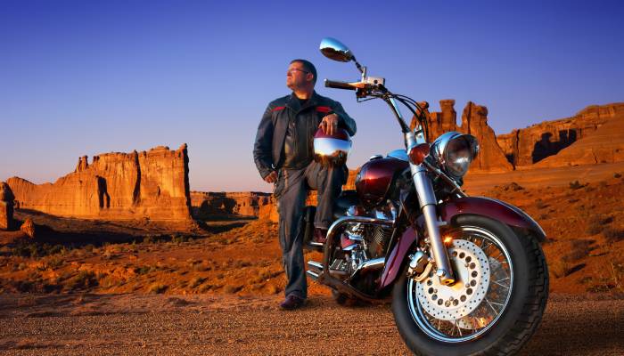 A motorcycle rider parked near the edge of the mountains. His helmet is on his knee as he rests against his bike.