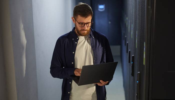 A man with a beard wearing glasses and a blue button-up over a white shirt stands and uses a laptop in a corridor.